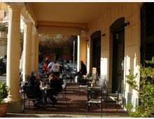 Portico di un edificio con grandi colonne chiare e soffitto alto, affacciato su una strada urbana. Sotto il portico c’è un’area esterna di caffè con tavolini e sedie in metallo, porte ad arco e alcune piante in vaso lungo il lato.