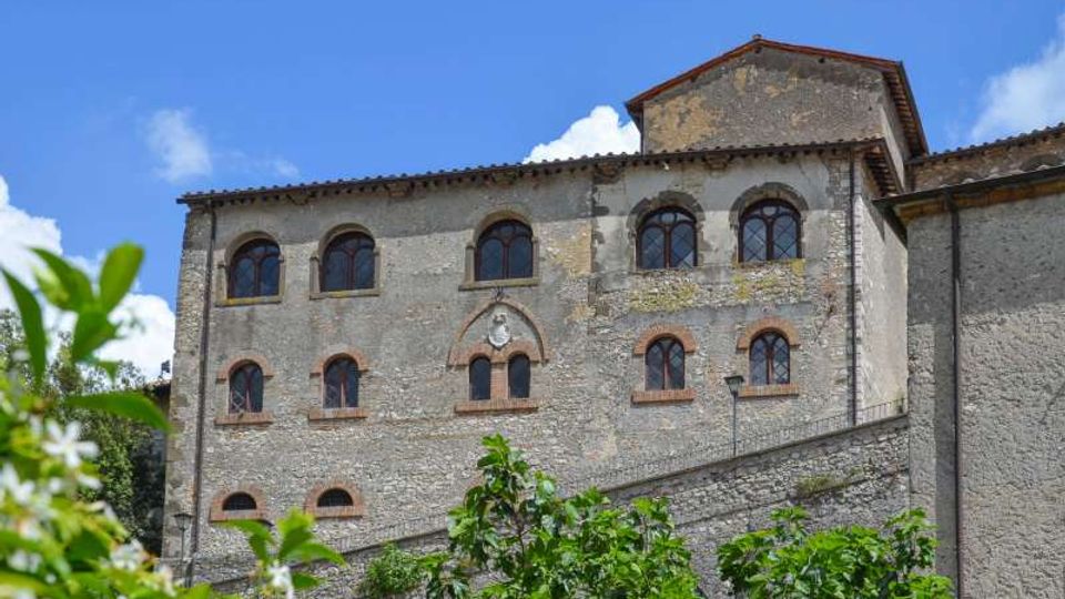 Facciata in pietra di un edificio storico a più piani, con numerose finestre ad arco e cornici in laterizio, sormontato da un tetto in tegole. In primo piano si vedono arbusti verdi e sullo sfondo un cielo azzurro con alcune nuvole.