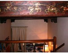 Interno di una casa con soppalco in legno e parapetto con colonnine, illuminato da una lampada da terra. In alto si vede una grande trave decorata con un dipinto di cavalieri, e sul fondo una tenda e scaffali con libri.