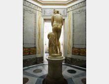 Interno di una sala museale classica con pavimento in marmo geometrico e pareti decorate, inquadrata verso una porta luminosa. Al centro, su un piedistallo, è esposta una statua in marmo a figura nuda vista di spalle, con drappo scolpito accanto.