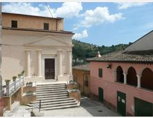 Piccola piazza di un borgo con una chiesa dalla facciata chiara, portale centrale e scalinata in pietra con ringhiere e fioriere. A destra si vede un edificio rosa con loggiato ad archi e tetto in coppi, sullo sfondo colline verdi sotto un cielo parzialmente nuvoloso.