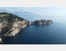 Veduta costiera con alte scogliere rocciose che scendono nel mare calmo e, al largo, una fortezza in pietra su un isolotto collegato alla terraferma. Sullo sfondo si intravedono montagne e una costa lontana sotto un cielo limpido.