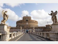 Veduta frontale di Castel Sant’Angelo con la sua struttura cilindrica in mattoni e le mura merlate, sullo sfondo di un cielo parzialmente nuvoloso. In primo piano il ponte con balaustre in pietra e statue di angeli scolpite lungo i lati conduce all’ingresso del castello.