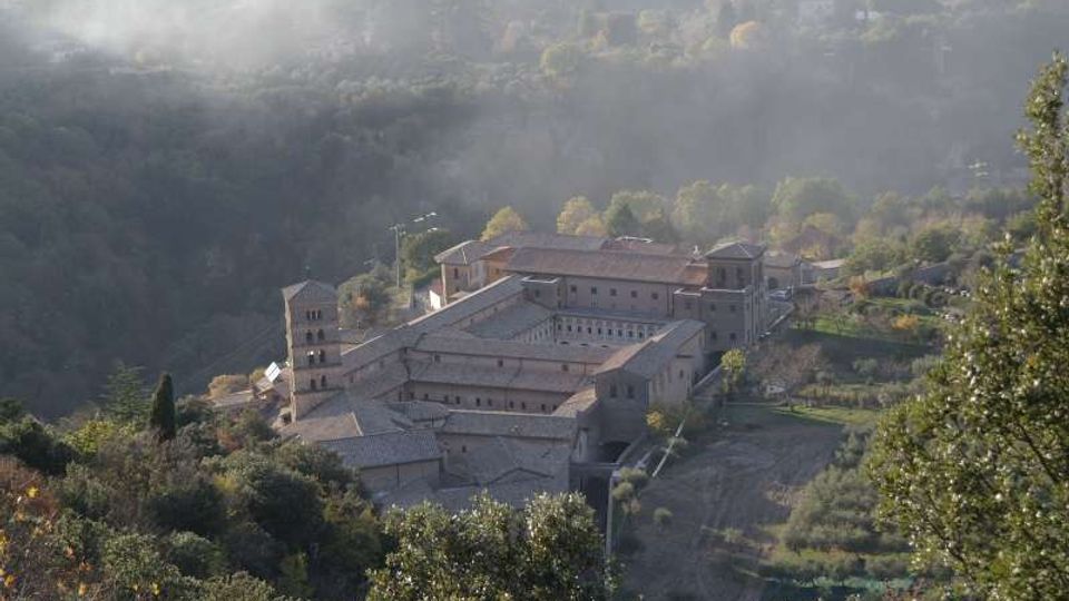Vista dall’alto di un grande complesso monastico in pietra con campanile e tetti in tegole, disposto attorno a un chiostro interno. L’edificio è circondato da boschi e campi su un pendio collinare, con foschia nelle colline sullo sfondo.