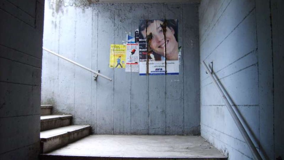 Sottopassaggio o scala di cemento grezzo con gradini e corrimano laterali, illuminato da una luce fredda dall’alto. Sulla parete di fondo sono affissi diversi manifesti e locandine.