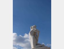 Statua in marmo di due figure nude abbracciate su un piedistallo, ripresa dal basso contro un cielo azzurro con alcune nuvole. Ambiente esterno con ampio sfondo di cielo.