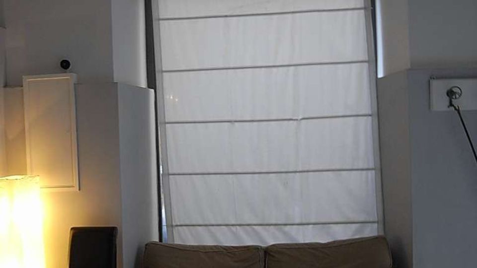 Interno di una stanza con grande finestra ad arco dotata di inferriata decorativa e tenda a rullo bianca. Sotto la finestra c’è un divano a due posti, con una sedia e una lampada da terra su pavimento in graniglia.