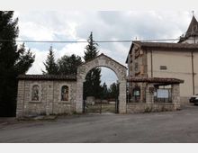 Ingresso in pietra di un complesso religioso con grande arco centrale e cancellata in ferro, affiancato da un muretto con nicchie decorate e da un edificio con tetto in coppi. Sullo sfondo si vedono alberi alti e cielo nuvoloso lungo una strada asfaltata.