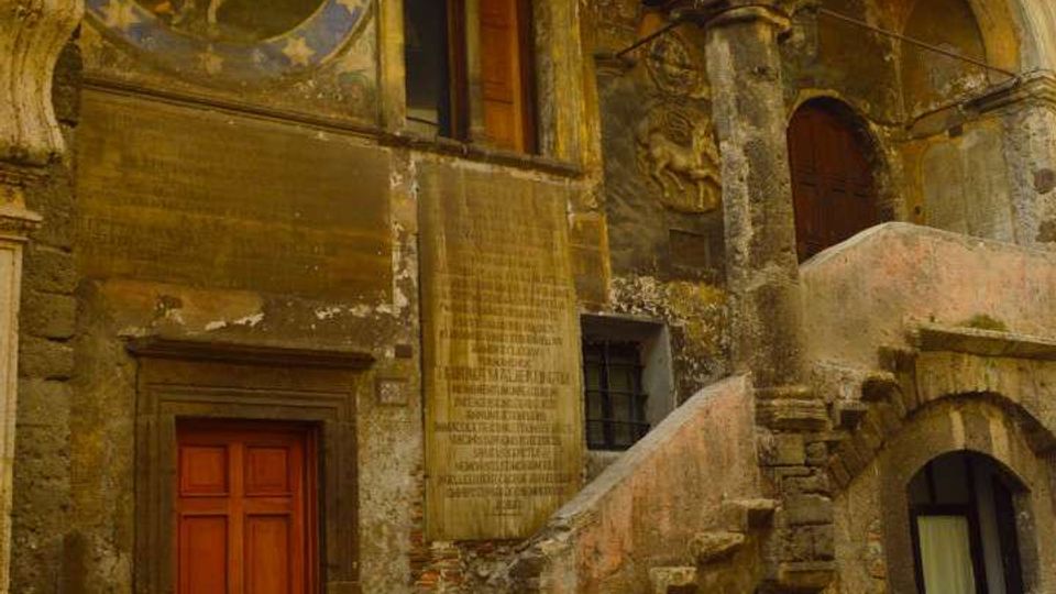 Facciata di un edificio storico in pietra con loggiato ad archi e colonne, affreschi murali consumati e vegetazione che spunta dalle cornici. Una scalinata esterna in muratura conduce al piano superiore con porte e finestre ad arco, affacciate su una strada lastricata.