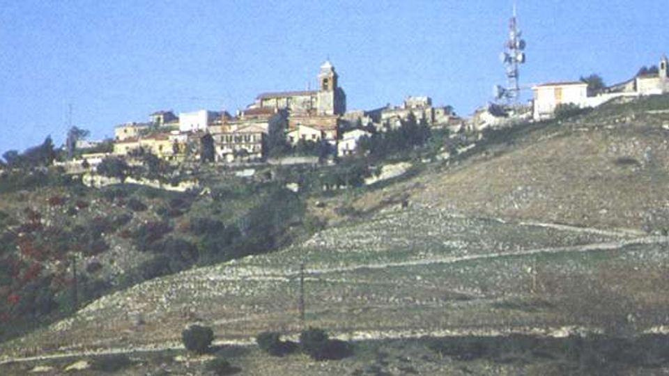Panorama di un piccolo borgo su un crinale collinare, con case compatte e una chiesa con campanile al centro. In primo piano si vede un pendio brullo con tracce di sentieri e, sulla destra, un’alta antenna di telecomunicazioni.