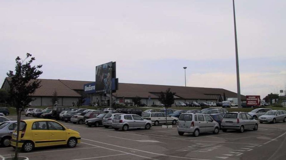 Ampio parcheggio asfaltato con numerose auto e segnaletica a terra, davanti a un grande edificio commerciale basso con tetto inclinato e insegna “Unieuro”. Sullo sfondo si vedono alti pali di illuminazione e alcune aiuole con piccoli alberi.