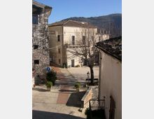 Piccola piazza di un borgo con edifici in pietra e intonaco dai tetti in coppi, affacciati su una strada lastricata. Al centro si vedono un albero spoglio, panchine, un lampione e alcune fioriere, con colline boscose sullo sfondo.