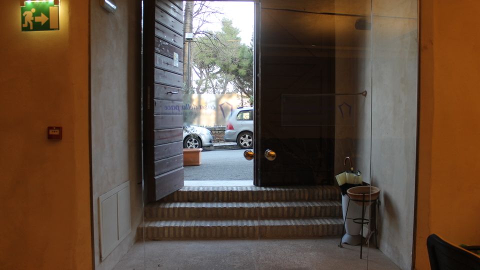Ingresso interno con porta vetrata e gradini in pietra che conducono all’uscita, affiancata da una parete in legno. All’esterno si vedono la strada con auto parcheggiate e alberi; all’interno è presente un cartello di uscita di emergenza e un portaombrelli.
