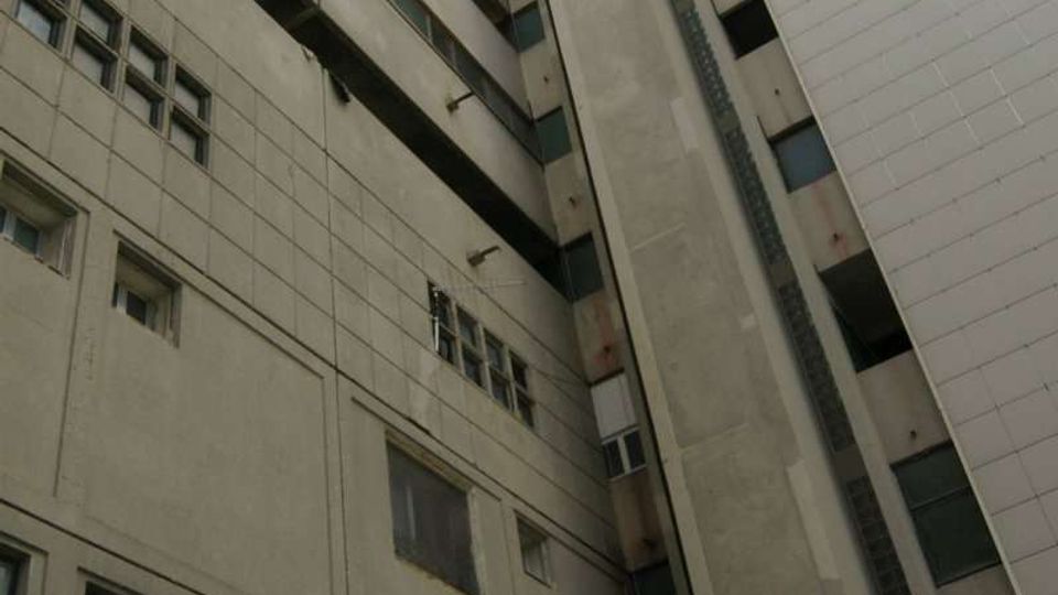 Veduta dal basso di un alto edificio multipiano in cemento, con facciate grigie e bianche e numerose finestre. Al centro si nota un grande vano verticale tipo torre tecnica o ascensore esterno che sale fino al tetto, con cielo visibile in alto tra le pareti.
