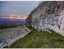 Spiazzo in pietra acciottolata accanto a un alto muro di fortificazione in pietra, con tratti di muratura antica e blocchi irregolari. Sullo sfondo si vede una vasta valle urbana al tramonto, con cielo nuvoloso e orizzonte montuoso.