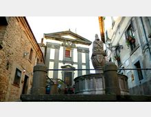 Piccola piazza tra edifici in pietra e intonaco, con una fontana circolare in primo piano sormontata da una scultura. Sullo sfondo si vede la facciata di una chiesa con frontone e lesene, raggiungibile tramite una breve scalinata.