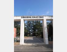 Ingresso di una caserma con grande portale rettangolare in cemento, con la scritta “CASERMA F.B. AZZANI” in alto. Oltre l’arco si vede un viale pavimentato che conduce a un’area interna alberata con alti pini e lampioni.