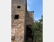 Facciata laterale di un edificio storico in pietra con finestre rettangolari e un passaggio ad arco che conduce a un corridoio ombreggiato. Vegetazione fitta sul lato destro e cielo azzurro sullo sfondo.