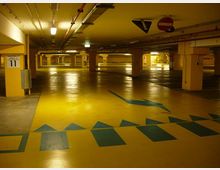 Parcheggio sotterraneo multipiano con pavimento giallo lucido e segnaletica direzionale blu, illuminato da lampade al neon. Pilastri numerati sostengono il soffitto basso con tubazioni a vista e cartelli stradali appesi.