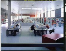 Ampia sala di biblioteca moderna con soffitto alto a pannelli e grandi finestre laterali che illuminano lo spazio. Sono presenti tavoli con postazioni computer, sedie e lunghe file di scaffali con libri disposti in modo ordinato.