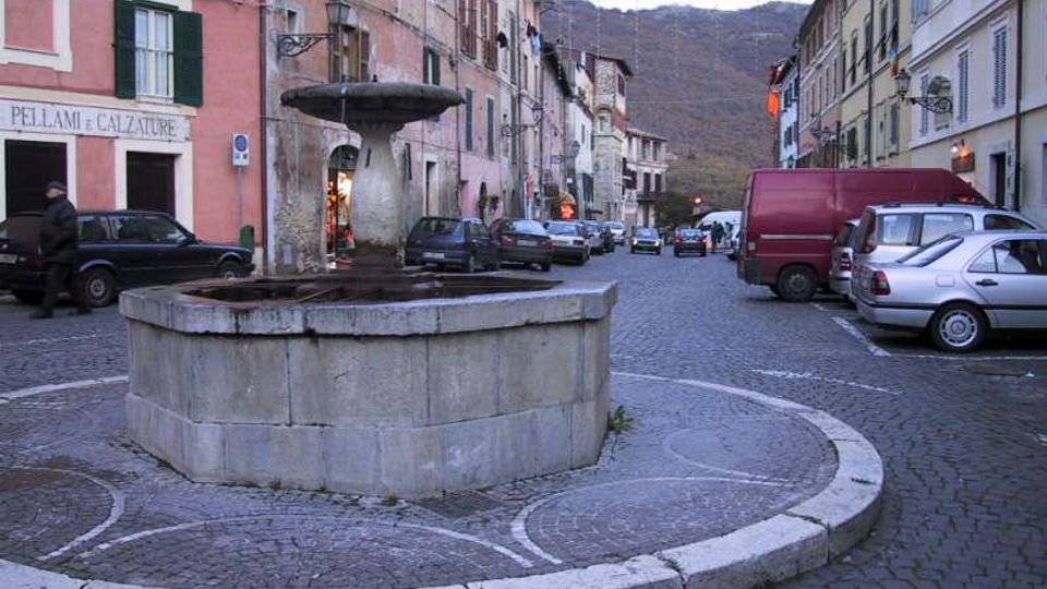 Fontana in pietra al centro di una piazzetta lastricata con pavé, circondata da edifici storici dai colori pastello e auto parcheggiate lungo la strada. Sullo sfondo si vedono colline o montagne oltre le case.
