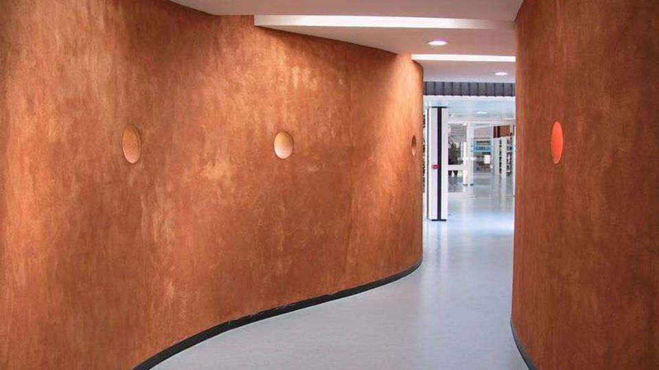 Corridoio interno moderno con pareti curve color terracotta e piccoli oblò circolari incassati, pavimento grigio chiaro e soffitto bianco con luci integrate. Il passaggio sinuoso conduce a un’area più ampia e luminosa visibile in fondo.
