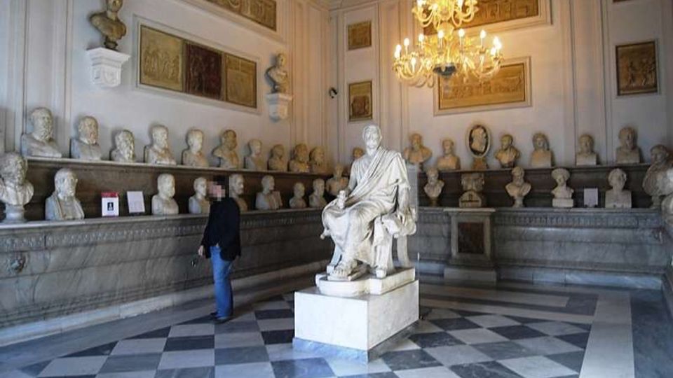 Sala museale neoclassica con pavimento a scacchi in marmo e pareti decorate con rilievi e pannelli, illuminata da un grande lampadario. Lungo il perimetro sono esposti numerosi busti in marmo su mensole, con una statua seduta su piedistallo al centro della stanza.