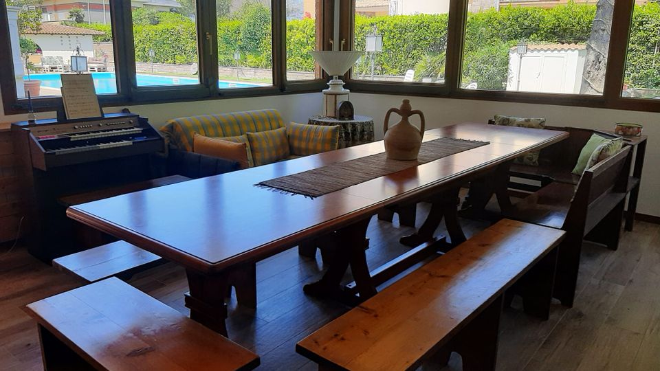 Sala da pranzo luminosa con grande tavolo in legno e panche, affiancata da divano e tastiera elettronica, circondata da ampie finestre angolari. Dalle vetrate si vede un giardino con siepi e una piscina all’esterno.