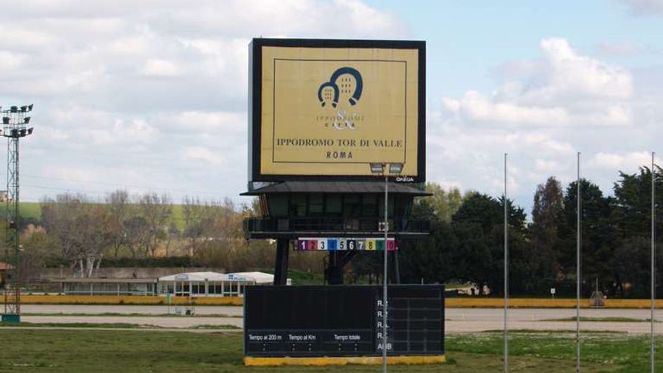 Grande tabellone segnapunti con schermo e pannelli informativi al centro dell’ippodromo Tor di Valle a Roma, circondato da piste e prati. Sullo sfondo si vedono alberi e cielo con nuvole.