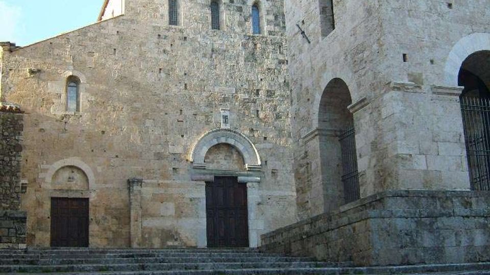 Facciata di una chiesa romanica in pietra chiara, con portali ad arco e piccole finestre, affacciata su un’ampia scalinata in pietra. A destra si vede una torre massiccia con grandi arcate e aperture strette sotto un cielo sereno.
