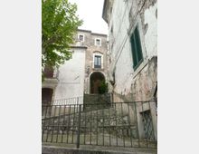 Scalinata in pietra con ringhiera metallica che sale tra edifici storici intonacati e in muratura, con arco di passaggio e balconcini. A sinistra si vede un albero e, sulla facciata a destra, una finestra con persiana verde.