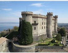 Veduta esterna di un grande castello in pietra con torri cilindriche merlate e mura alte, affacciato su una collina con giardini e un vialetto d’accesso. Sullo sfondo si vede un lago e un paesaggio collinare sotto un cielo sereno.