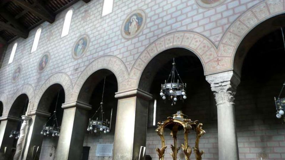 Interno di una chiesa con navata laterale scandita da grandi archi su colonne, soffitto ligneo e lampadari sospesi. Sulla parete in muratura sono presenti finestre alte e medaglioni decorativi, con un baldacchino dorato in primo piano.