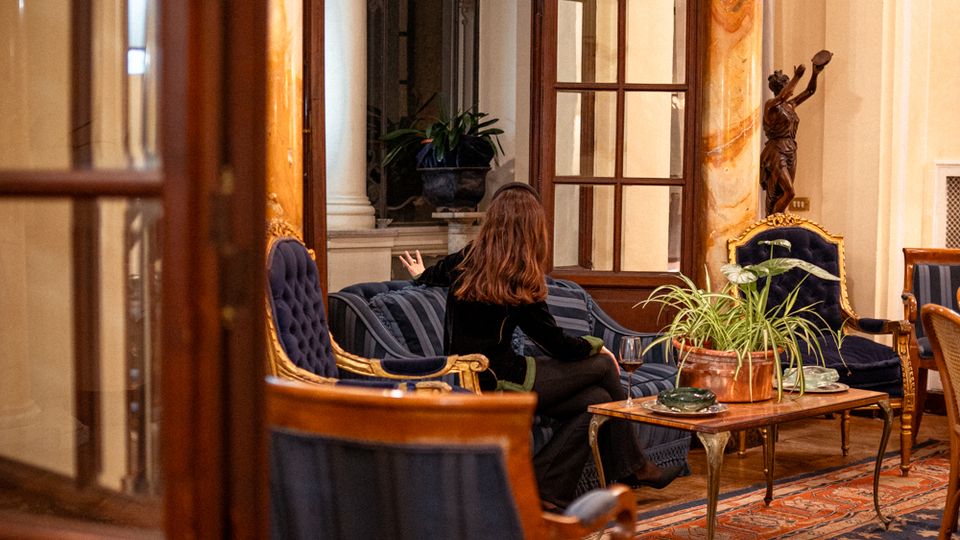 Salotto interno in stile classico con poltrone e divani imbottiti blu, tavolini bassi e tappeti decorati. L’ambiente è definito da alte colonne in marmo, grandi porte-finestre con telai in legno e una statua su piedistallo vicino alla parete.