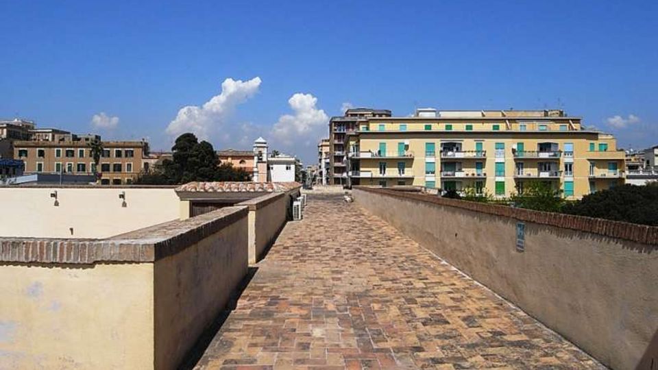 Camminamento sopraelevato con pavimentazione in mattoni e muretti laterali intonacati, che conduce verso edifici residenziali. Sullo sfondo si vedono palazzi a più piani e un cielo sereno con alcune nuvole.