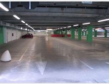Parcheggio sotterraneo con ampie corsie in cemento, illuminazione a neon e soffitto con tubazioni a vista. Pilastri verdi e segnaletica con numeri dei posti delimitano le aree di sosta.