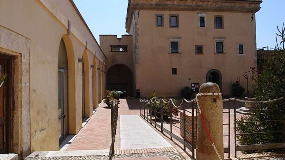 Cortile interno con pavimentazione in mattoni e percorso centrale, affiancato a sinistra da un porticato ad archi e di fronte da un edificio storico a più piani con finestre rettangolari. Sono presenti ringhiere e catene che delimitano le aiuole, con un grande pilastro in pietra in primo piano.