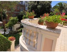Terrazza esterna con balaustra curva in pietra chiara e vasi di terracotta con fiori rossi, affacciata su un giardino verde sottostante. Sullo sfondo si vedono alberi e palme e un edificio residenziale con balconi.