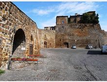 Piazza lastricata in pietra ai piedi di una fortificazione medievale, con alte mura in pietra e un grande arco sul lato sinistro. Sullo sfondo si vede il bastione con edifici in pietra in cima e alcune auto parcheggiate lungo il muro.