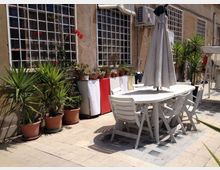 Terrazza esterna pavimentata accanto a un edificio con grandi finestre a griglia metallica, con un tavolo e sedie bianchi sotto un ombrellone chiuso. Lungo la parete ci sono numerosi vasi con piante e una fila di piccoli contenitori, oltre a un’unità di condizionamento fissata in alto.