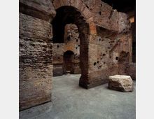 Interno di rovine in mattoni con grandi archi a tutto sesto e pareti irregolari segnate da fori e tracce di muratura antica. Sul pavimento in cemento si vede un grande blocco di pietra squadrato accanto a un corridoio che conduce ad altri passaggi ad arco.