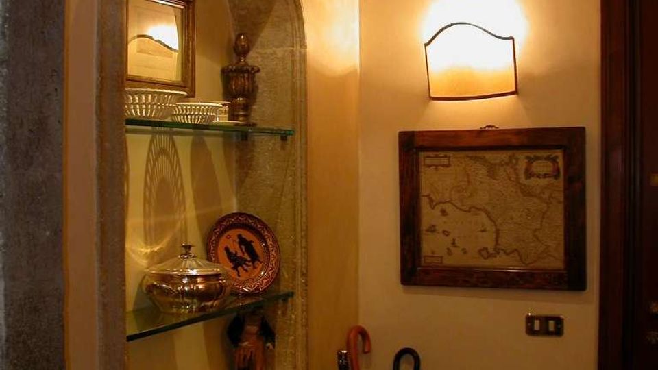 Ingresso interno con nicchia ad arco in pietra e mensole in vetro che espongono oggetti decorativi. Pareti color crema con applique a parete, quadro con mappa incorniciata e porta scura; a terra un portaombrelli con ombrelli.
