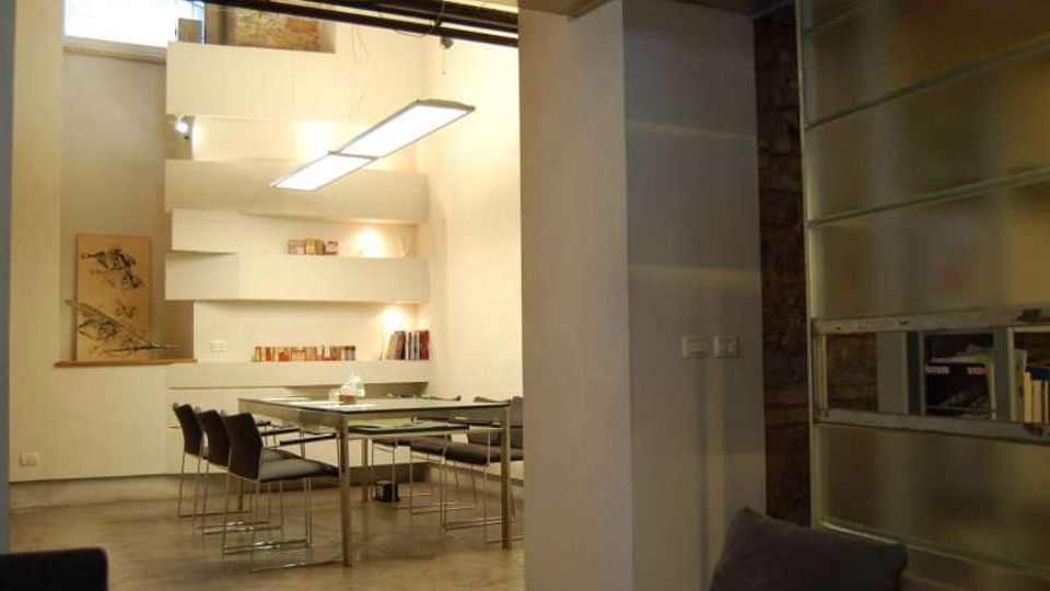 Interno di un open space moderno con pavimento in cemento, tavolo da pranzo in vetro e sedie scure, illuminato da lampade rettangolari sospese. Sul fondo ci sono nicchie e mensole bianche con libri e una piccola finestra alta, mentre a destra si vede una parete vetrata satinata e un grande pilastro.