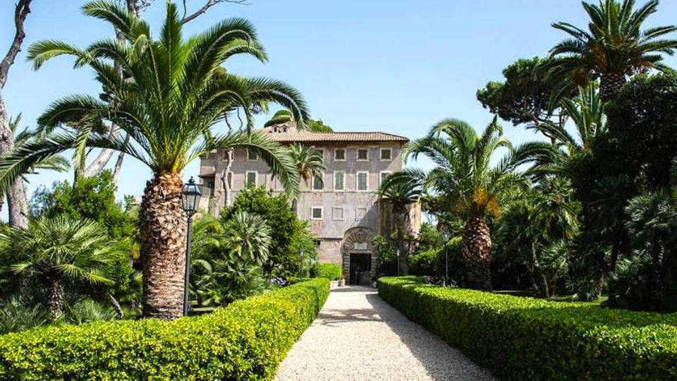 Viale di ghiaia in un giardino formale, fiancheggiato da siepi potate e alte palme, che conduce a una villa storica con facciata in pietra, finestre regolari e portale ad arco. Sullo sfondo si vede il cielo sereno e una vegetazione mediterranea densa ai lati del percorso.
