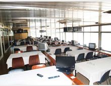 Ampia sala interna con file di tavoli rettangolari coperti da tovaglie bianche e sedie, con numerosi monitor e televisori distribuiti sui tavoli e lungo le pareti. Grandi finestre laterali illuminano l’ambiente, con soffitto metallico e ventilatori circolari.