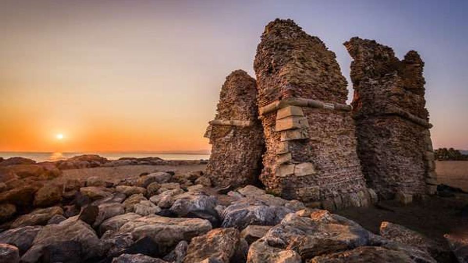 Rovine in pietra di una struttura costiera con grandi muri parzialmente crollati, affacciata su una spiaggia rocciosa. In primo piano massi e scogli, con il mare e il sole basso all’orizzonte sullo sfondo.