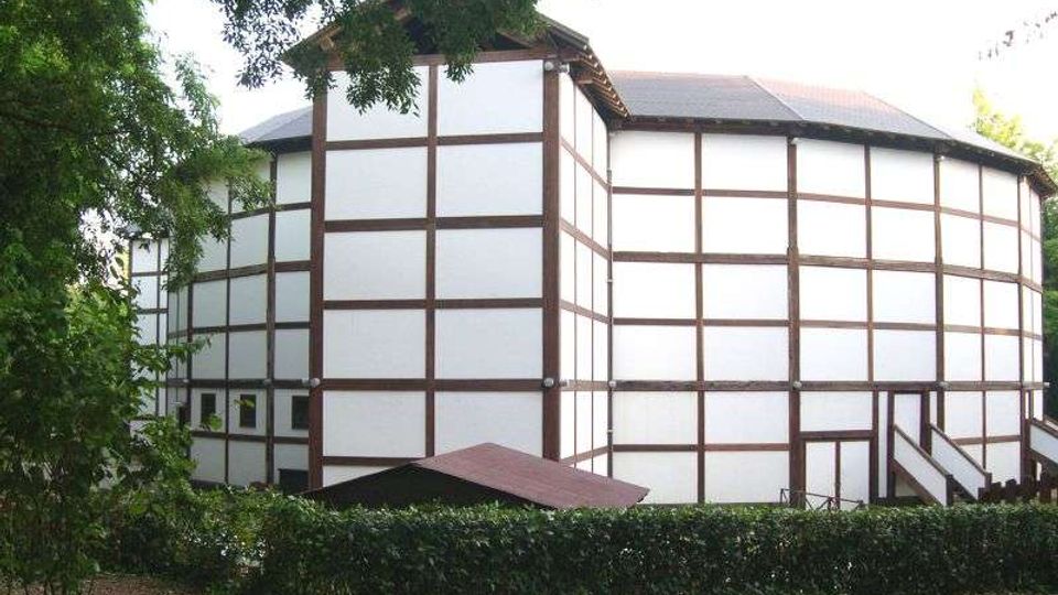 Esterno di un teatro circolare in stile Tudor, con pareti bianche a graticcio di travi scure e tetto basso, circondato da alberi e siepi. Una piccola scala laterale e una tettoia d’ingresso sono visibili vicino alla base dell’edificio.