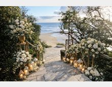 Scalinata in pietra che scende attraverso vegetazione costiera verso una spiaggia sabbiosa e il mare, con ringhiere in legno ai lati. Lungo i gradini sono disposte lanterne con candele e grandi composizioni floreali bianche su supporti dorati e a terra.