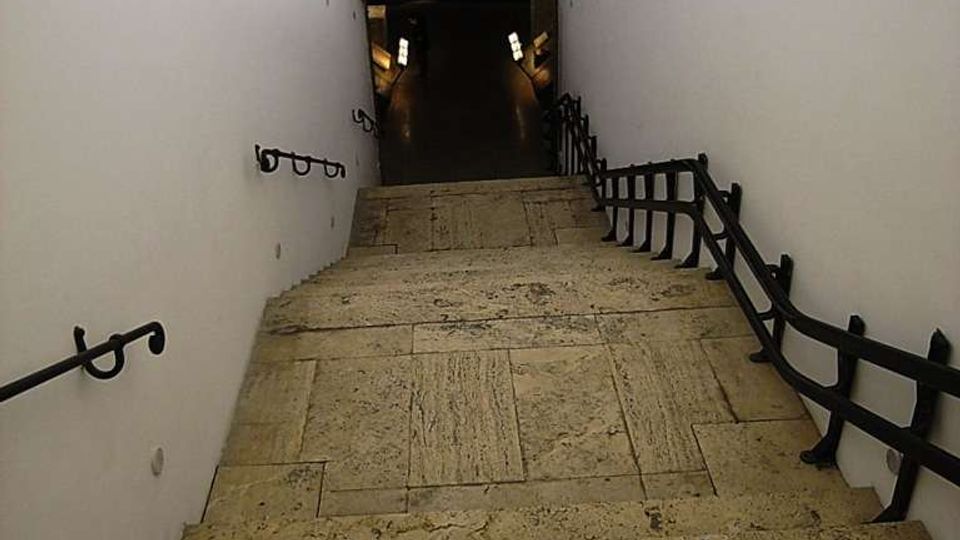 Scala in pietra che scende in un corridoio sotterraneo con pareti bianche e soffitto a volta. Corrimano e ringhiere nere lungo i lati conducono a un arco che apre su uno spazio più buio illuminato da piccole luci.