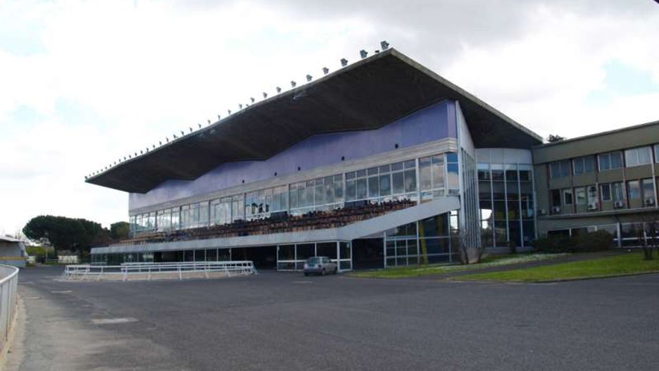 Vista esterna di una tribuna di ippodromo con grande tetto sporgente e facciata vetrata su più livelli, con rampa laterale e ingresso al piano terra. Davanti si estende una vasta area asfaltata, con prato e alcuni edifici annessi sul lato destro.